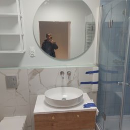 Usługi ogólnobudowlane Marcin Łobocki - Wykończona łazienka z okrągłym lustrem, białą umywalką nablatową na drewnianej szafce, toaletą i kabiną prysznicową z niebieskimi płytkami; widoczne zabezpieczenia taśmą malarską.