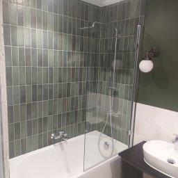 Usługi ogólnobudowlane Marcin Łobocki - Nowoczesna łazienka z wanną, szklanym parawanem nawannowym, deszczownicą i umywalką nablatową na ciemnym blacie, wykończona pionowymi, zielonymi płytkami.