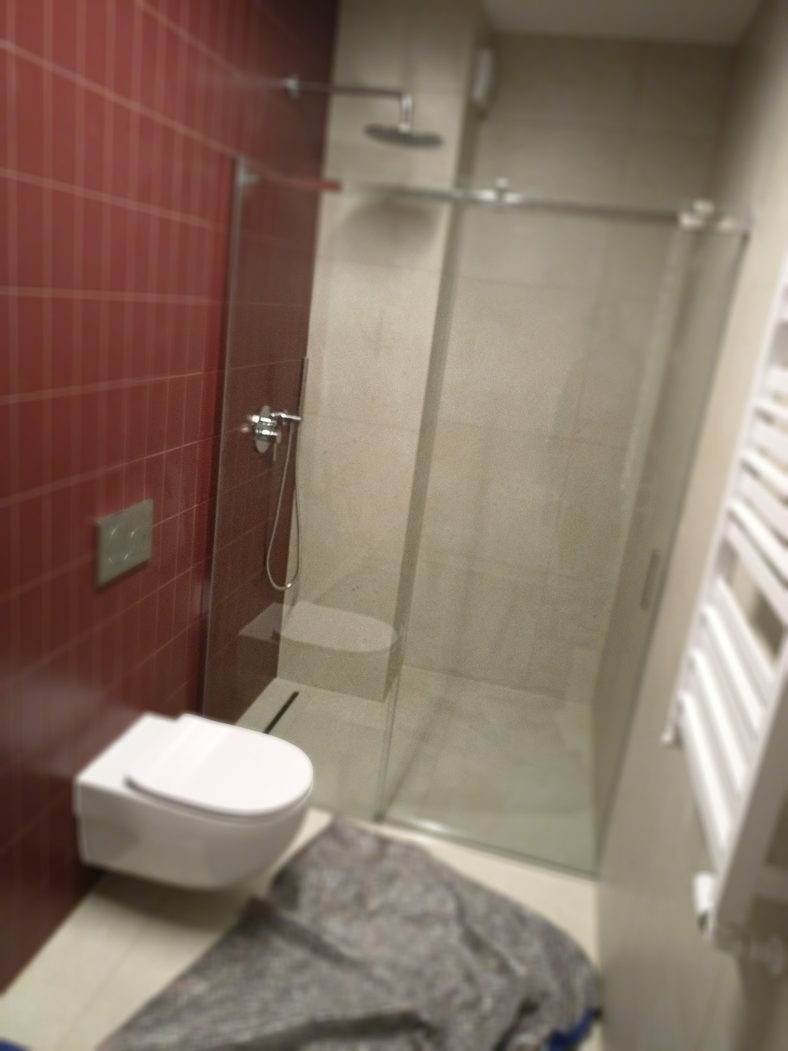 Nowoczesna łazienka z wiszącą toaletą, kabiną prysznicową ze szklanymi drzwiami i grzejnikiem drabinkowym, wykończona bordowymi płytkami na jednej ścianie.