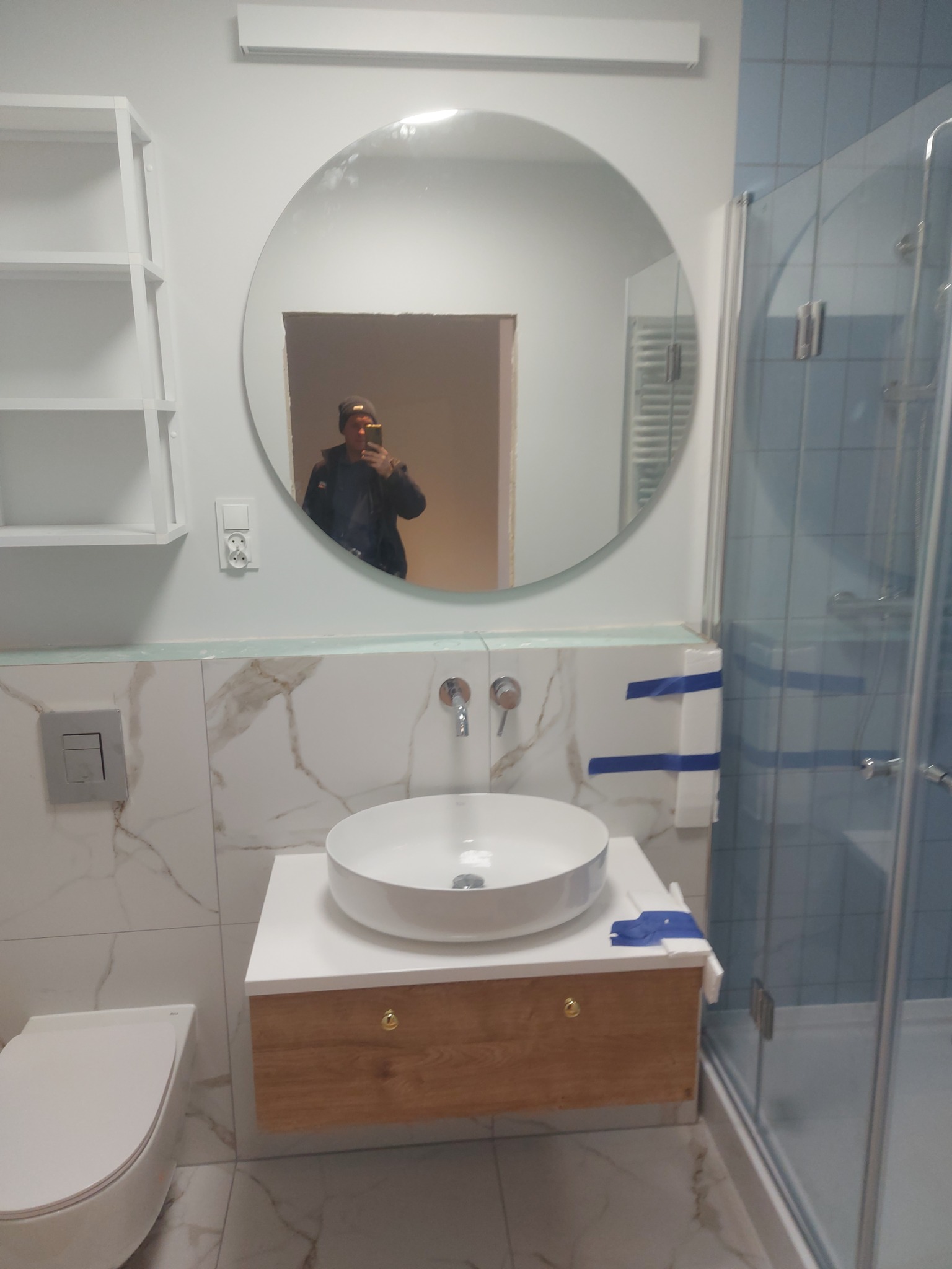 Wykończona łazienka z okrągłym lustrem, białą umywalką nablatową na drewnianej szafce, toaletą i kabiną prysznicową z niebieskimi płytkami; widoczne zabezpieczenia taśmą malarską.