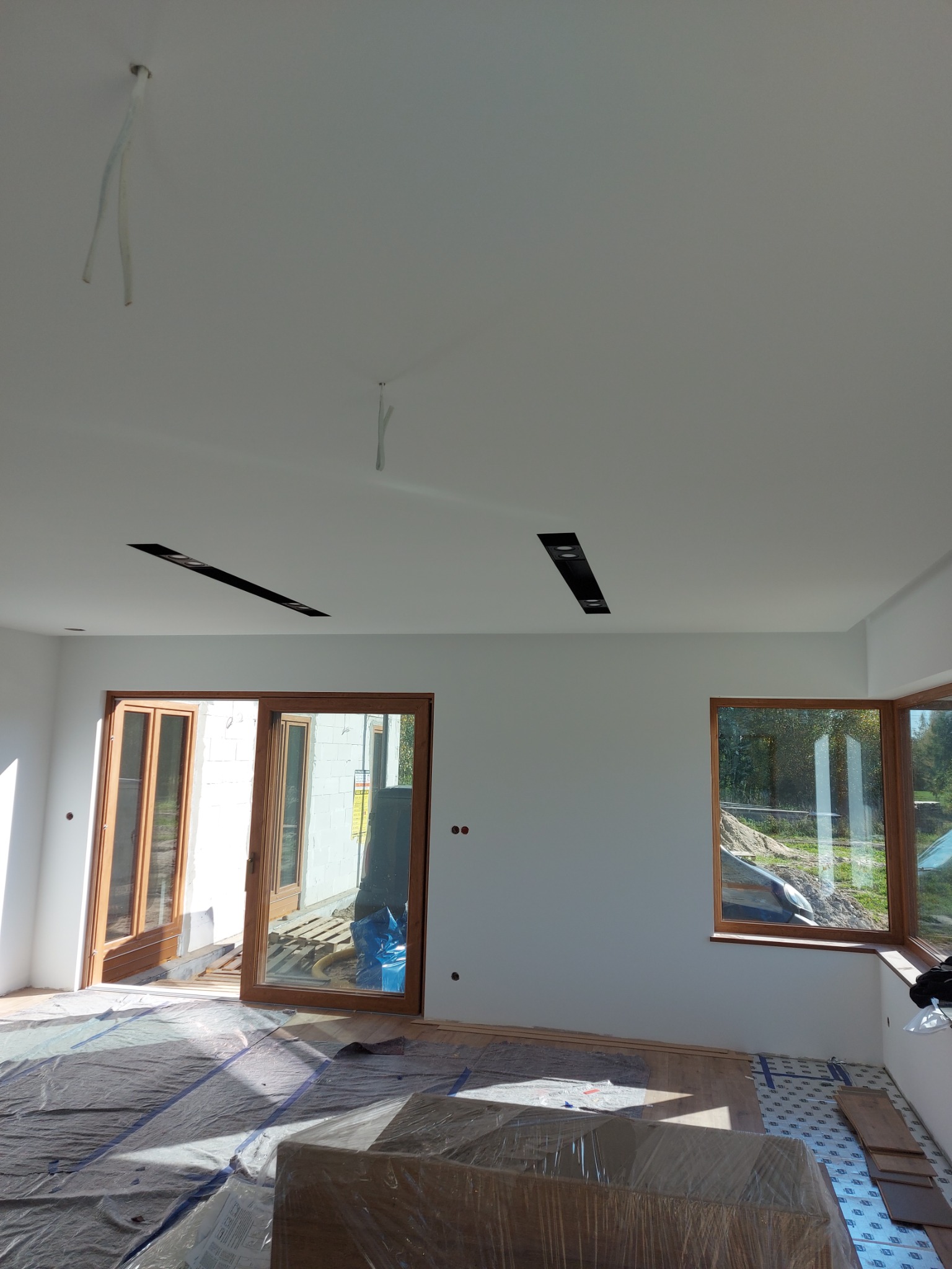 Wnętrze w trakcie wykańczania: białe ściany, przygotowane do montażu panele podłogowe zabezpieczone folią, widoczne okna i drzwi balkonowe z drewnianymi ramami oraz częściowo zamontowane...