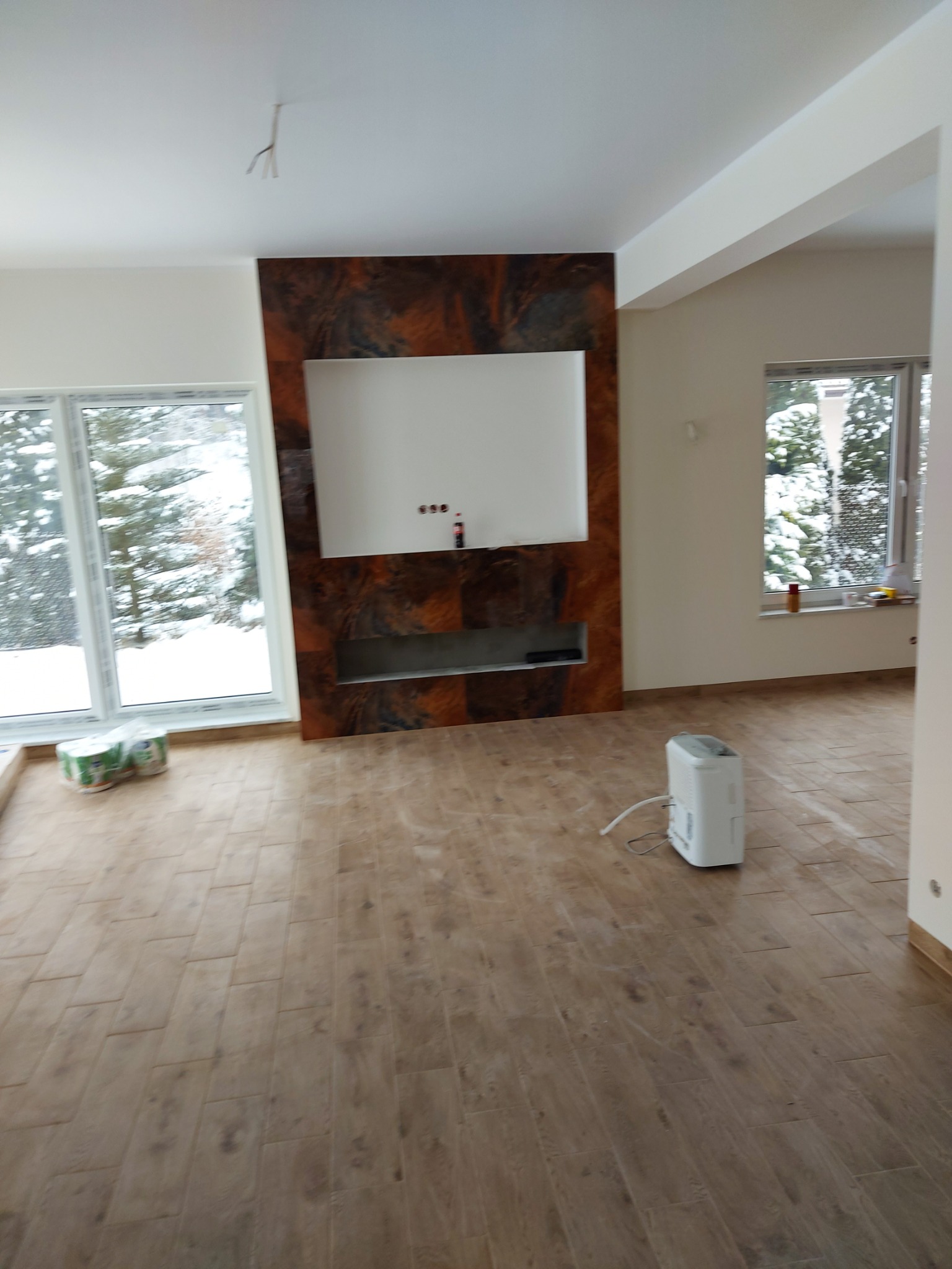 Ułożone panele podłogowe w jasnym salonie z dekoracyjną ścianą, widoczny osuszacz powietrza i okna z zimowym krajobrazem.