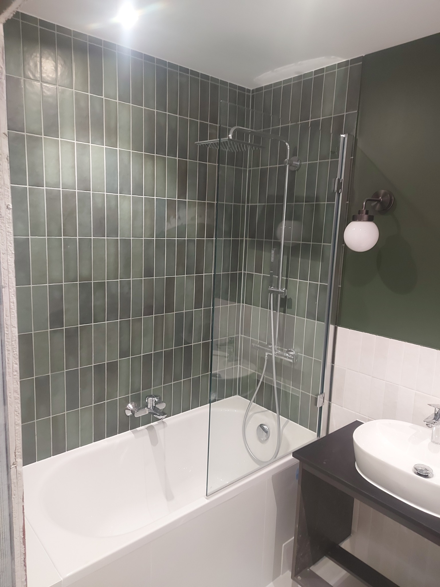 Nowoczesna łazienka z wanną, szklanym parawanem nawannowym, deszczownicą i umywalką nablatową na ciemnym blacie, wykończona pionowymi, zielonymi płytkami.