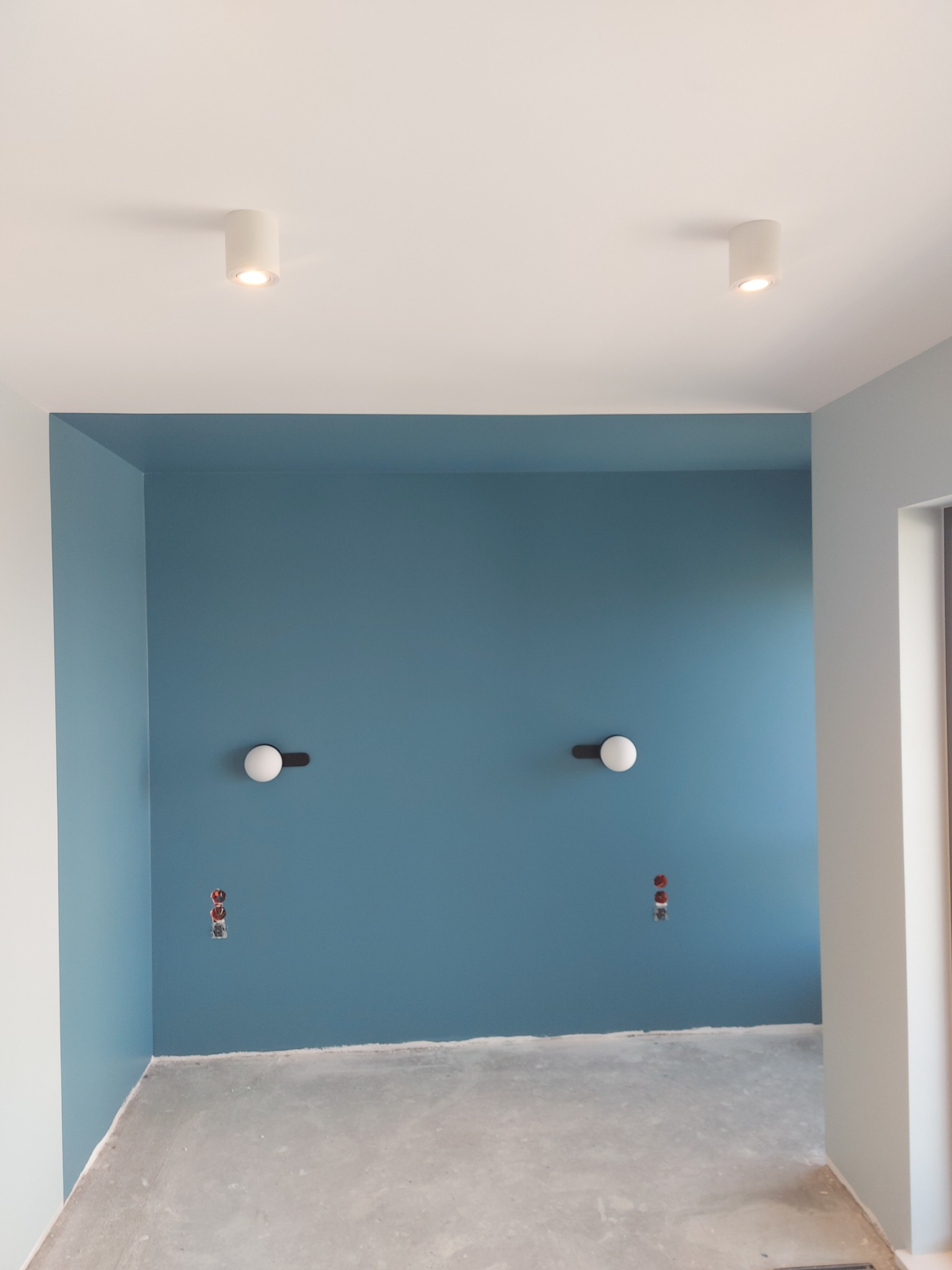 Świeżo pomalowane wnętrze z błękitnymi ścianami, widoczne dwa okrągłe kinkiety i puszki elektryczne, sufit z dwoma punktowymi lampami, surowa podłoga przed wykończeniem.