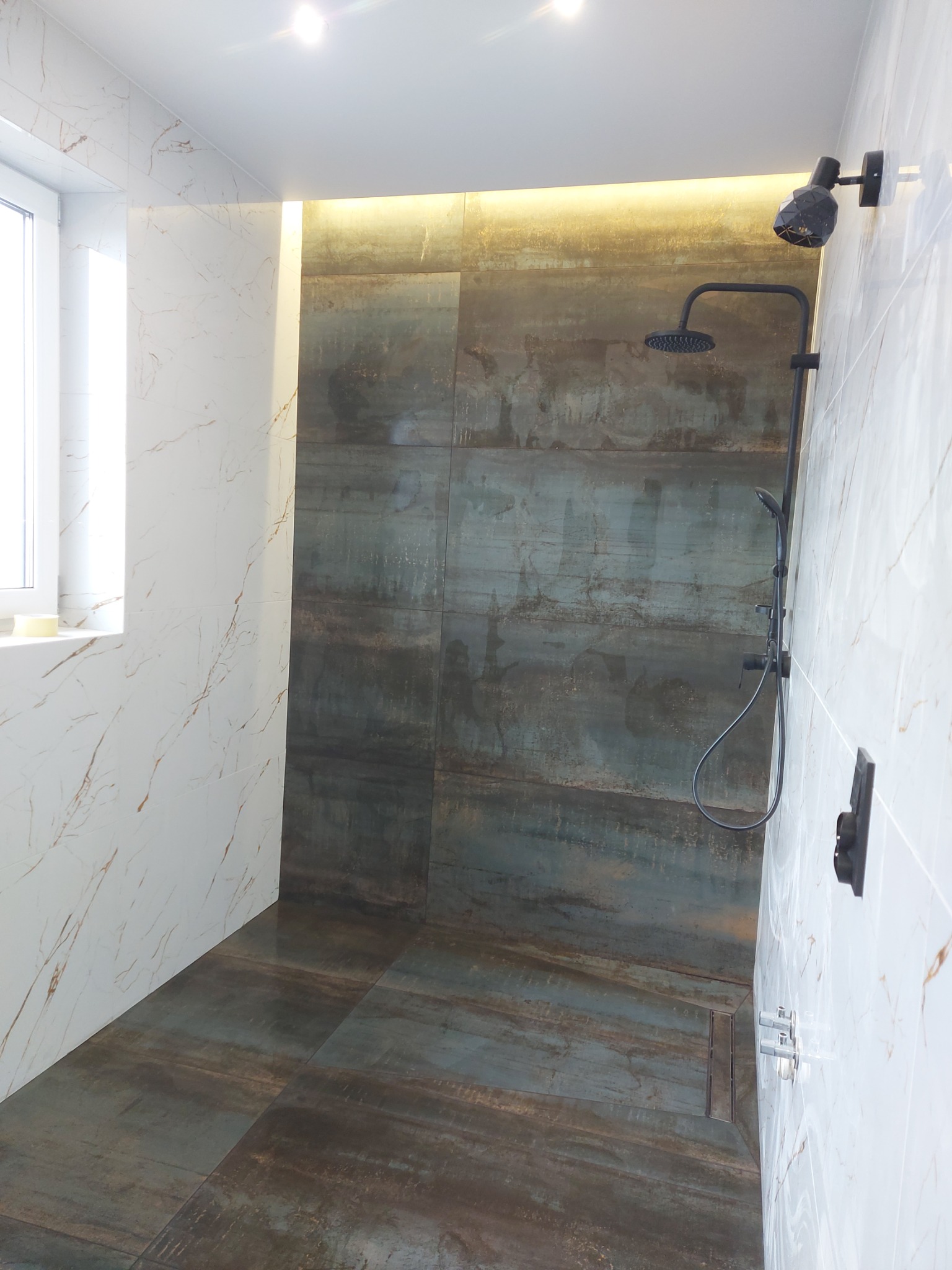 Nowoczesna kabina prysznicowa z czarną armaturą, wyłożona płytkami imitującymi marmur i metal, oświetlona punktowo.