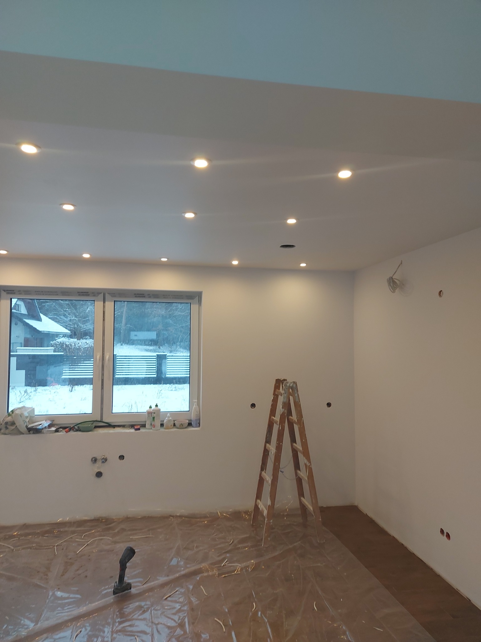 Wnętrze pokoju w trakcie prac wykończeniowych: pomalowane ściany na biało, zamontowane oświetlenie sufitowe, drewniana drabina, okno z widokiem na zimowy krajobraz, podłoga zabezpieczona folią...