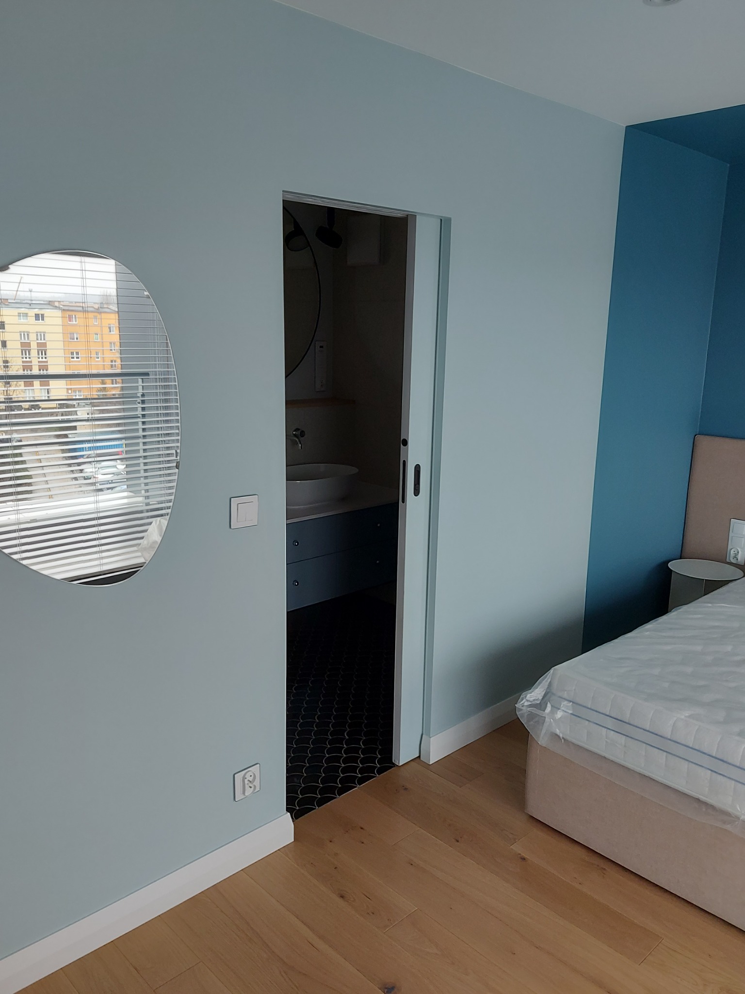 Nowoczesne wnętrze sypialni z błękitnymi ścianami, drewnianą podłogą i owalnym lustrem odbijającym widok z okna, z widocznym wejściem do łazienki z umywalką na szafce.