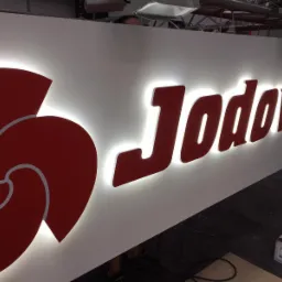 Podświetlane, przestrzenne litery i logo w kolorze czerwonym na białym tle, tworzące szyld 'Jodovit', z widocznymi elementami montażowymi pod sufitem i opakowaniem materiałów w tle.