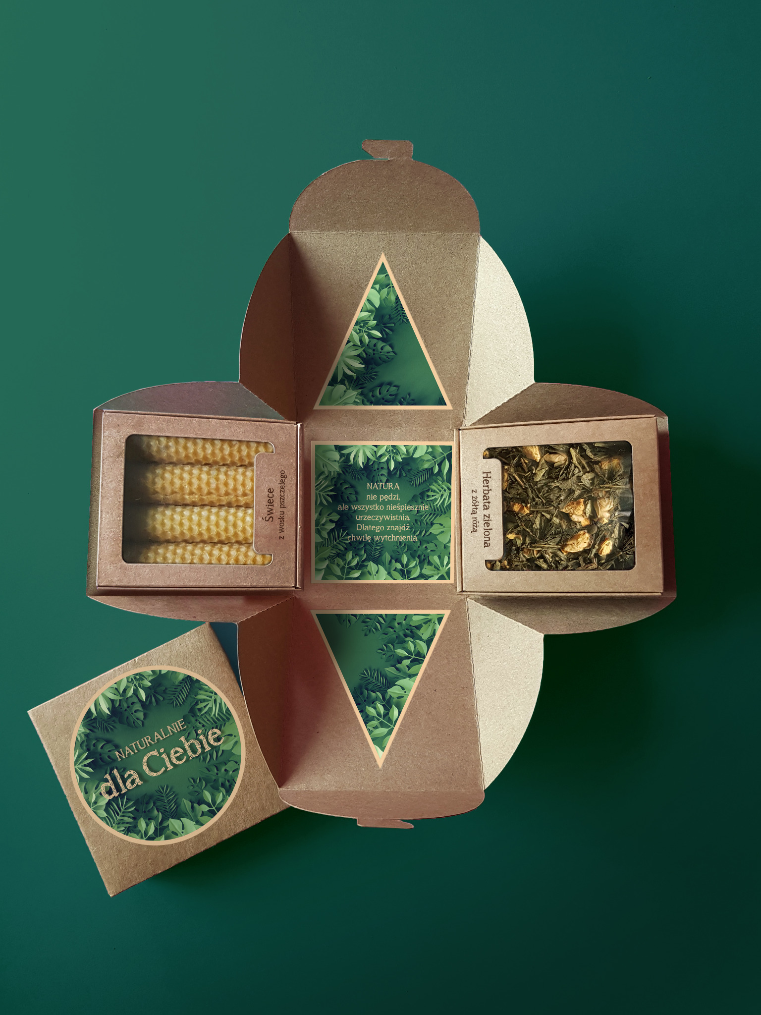 Otwarty, ekologiczny kartonowy upominek z zawartością: świece z wosku pszczelego, zielona herbata z żółtą różą oraz kartka z napisem 'Naturalnie dla Ciebie' i motywem roślinnym na ciemnozielonym tle.