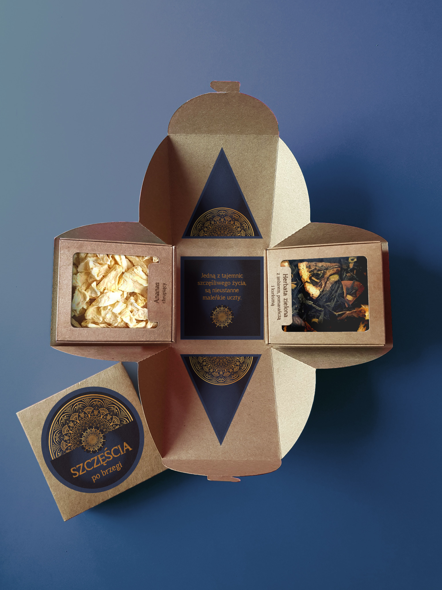 Otwarty, kwadratowy kartonowy box prezentowy z trzema przegródkami zawierającymi: suszonego ananasa, zieloną herbatę z imbirem, pomarańczą i kurkumą, oraz tekst z życzeniami, na ciemnoniebieskim tle.