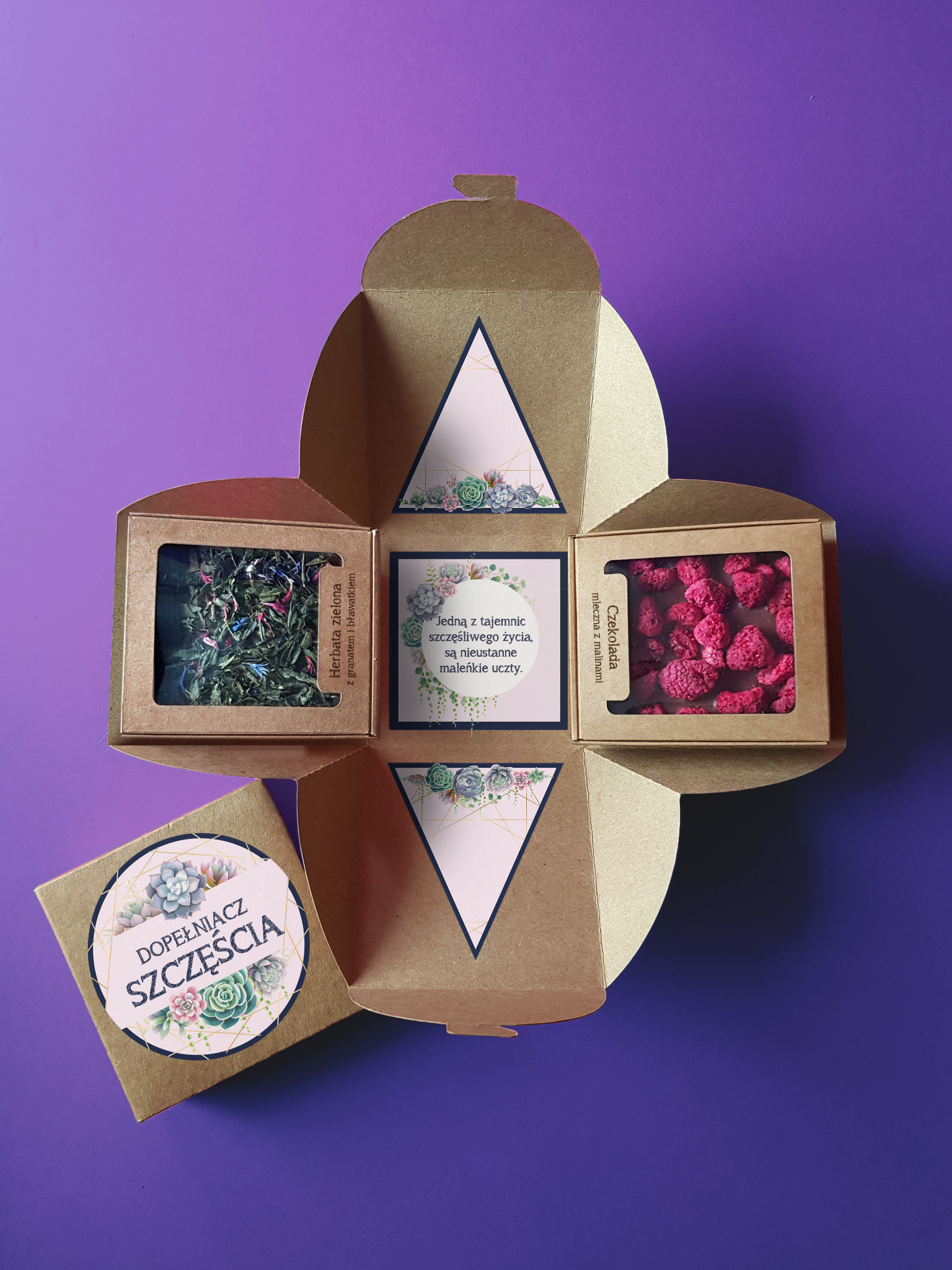 Otwarty, kwadratowy, złoty kartonik z czterema przegródkami, zawierający herbatę zieloną z granatem i bławatkiem, czekoladę mleczną z malinami oraz kartkę z sentencją, na fioletowym tle.