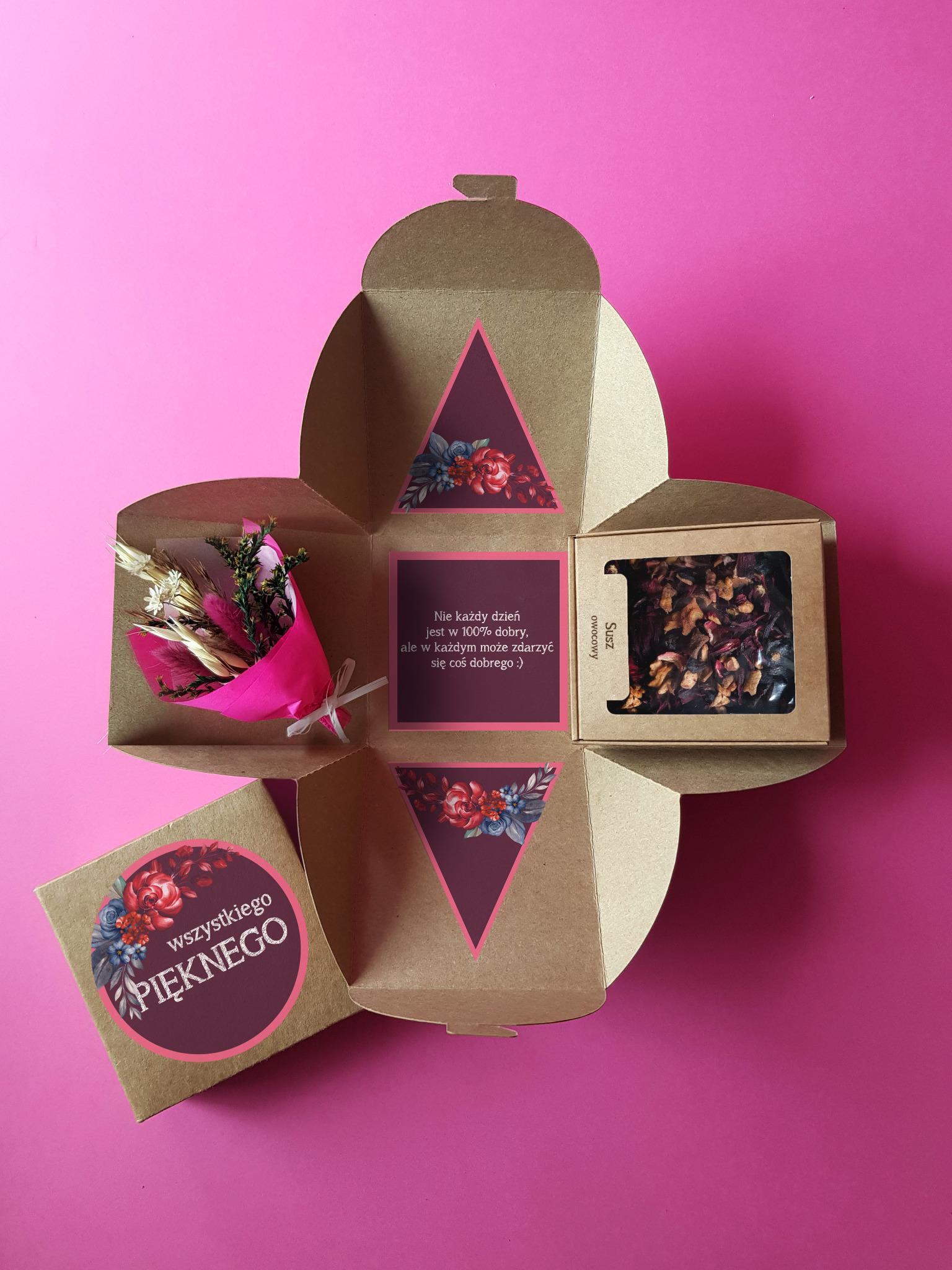 Otwarty, tekturowy, kwadratowy box z przegródkami na różowe tło. W jednej przegródce mały bukiecik z suszonych kwiatów w różowym papierze, w innej susz owocowy w małym, tekturowym pudełku...