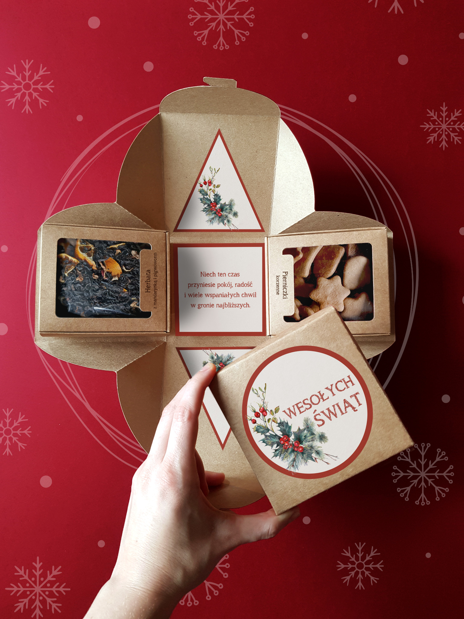 Otwarty, kwadratowy, tekturowy zestaw prezentowy z herbatą, pierniczkami i życzeniami świątecznymi, trzymany przez kobiecą dłoń na czerwonym tle z motywem śnieżynek.