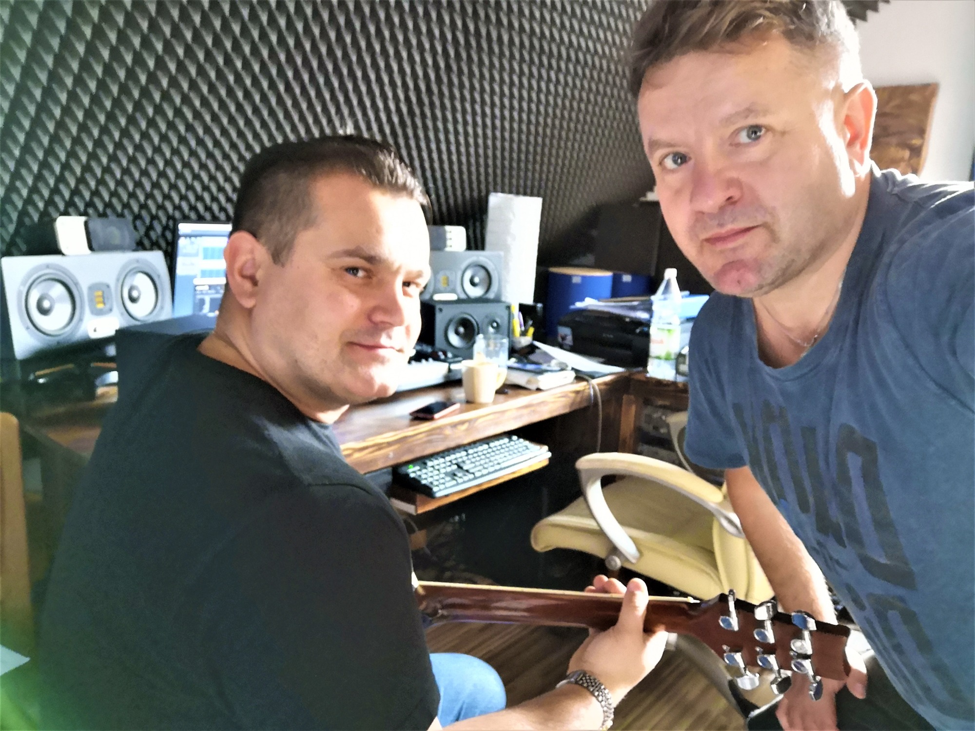 Dwóch mężczyzn w pomieszczeniu z panelami akustycznymi, jeden trzyma gitarę, widoczne monitory studyjne i klawiatura.