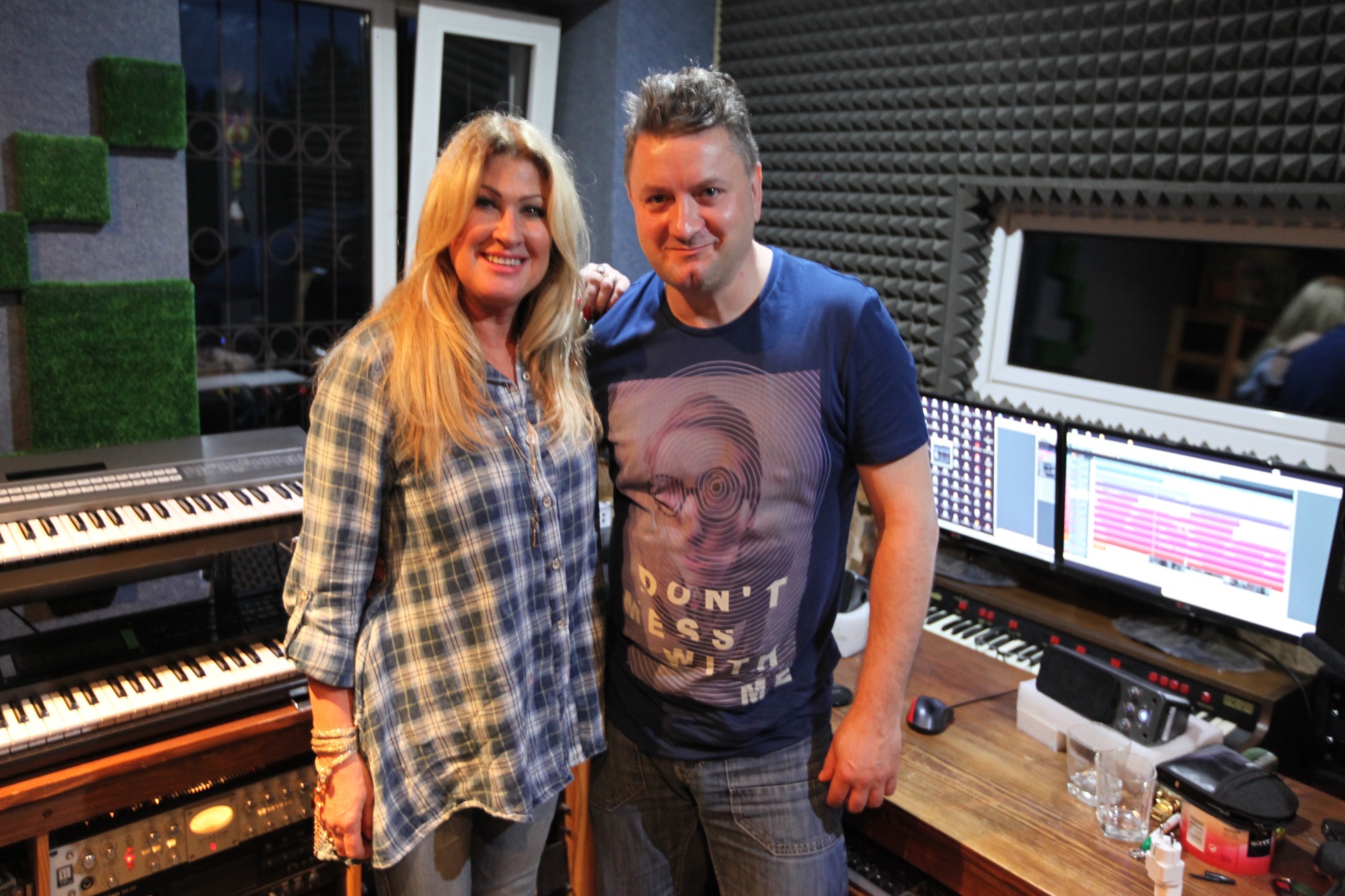 Kobieta i mężczyzna uśmiechają się w pomieszczeniu z pianinem, dwoma monitorami z oprogramowaniem do edycji audio oraz ścianą wytłumiającą dźwięk.
