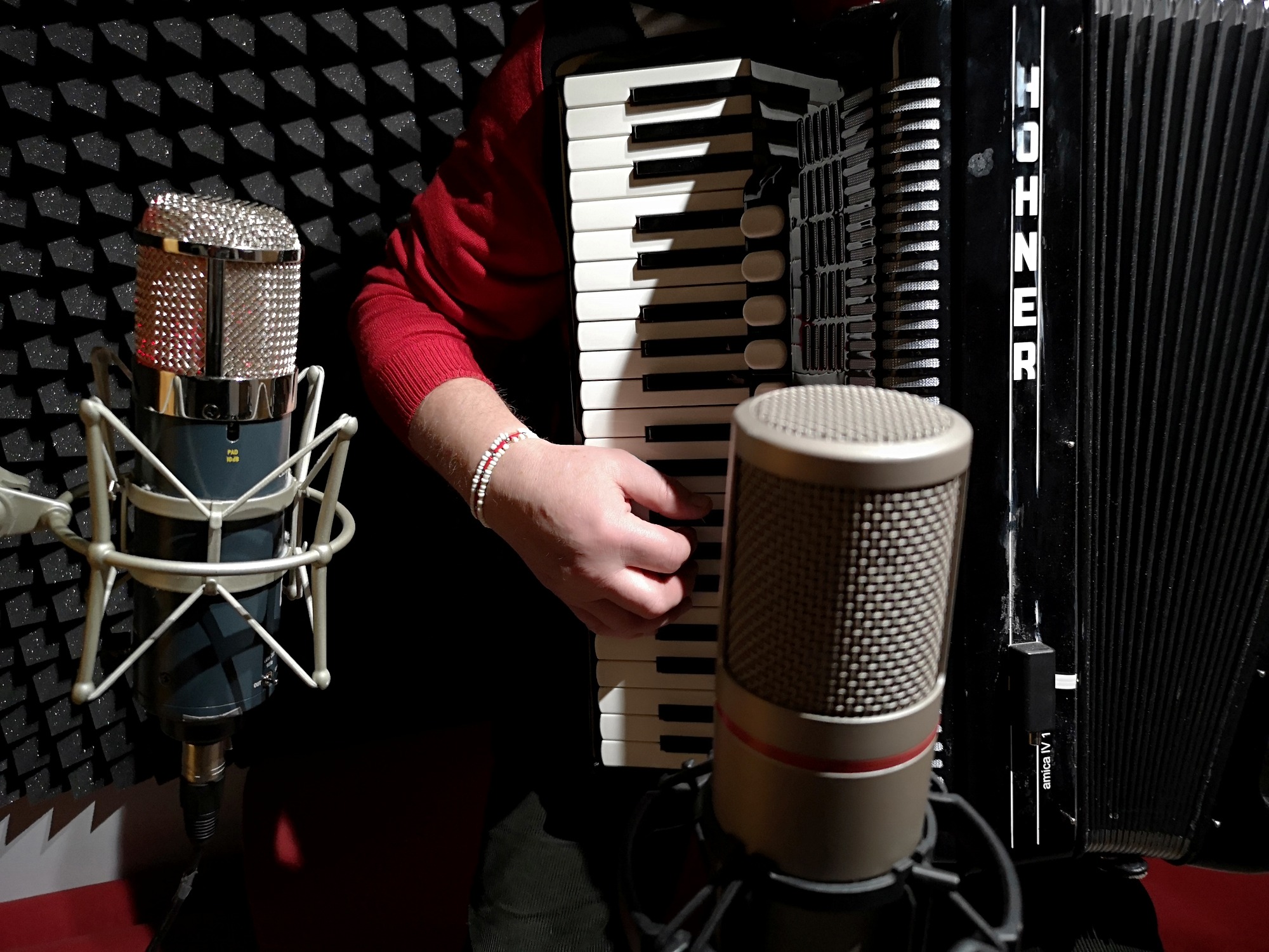 Osoba w czerwonym swetrze gra na akordeonie Hohner Amica IV 1 w pomieszczeniu z akustycznymi panelami, z dwoma mikrofonami studyjnymi.