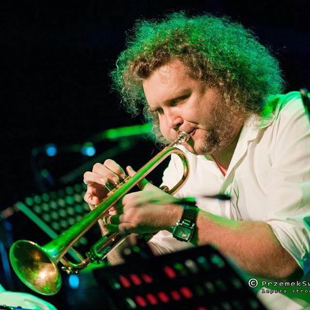 Muzyk grający na trąbce na scenie, oświetlony zielonym światłem, z widocznymi nutami i zegarkiem na ręku.