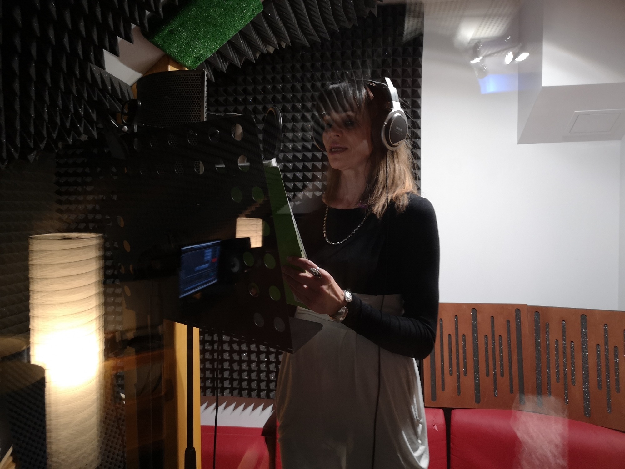 Kobieta w słuchawkach nagrywa tekst, czytając z zielonej kartki na statywie w wyciszonym pomieszczeniu z panelami akustycznymi.