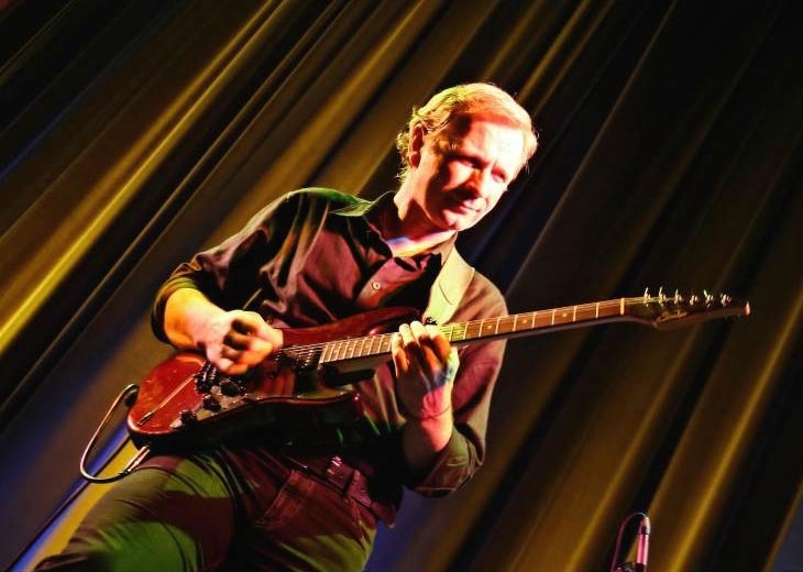 Gitarzysta grający na elektrycznej gitarze na scenie z żółto-czarnym draperią w tle.