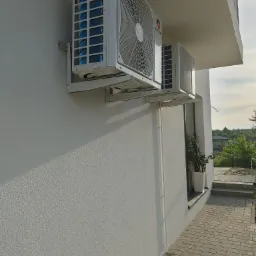 Dwa białe klimatyzatory marki Gree zamontowane na białej ścianie budynku z kostki brukowej w tle.