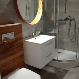 Nowoczesna łazienka z okrągłym lustrem w drewnianej ramie, białą umywalką z szafką, wiszącą toaletą i kabiną prysznicową ze srebrnym zestawem natryskowym na tle szarych płytek.