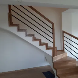 Nowoczesne schody z drewna z czarnymi, metalowymi prętami poręczy, oświetlone naturalnym światłem, widok z dołu na jasne wnętrze.