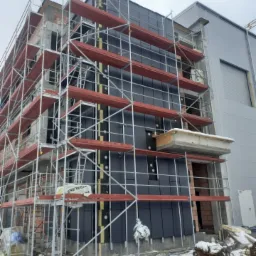 Budynek biurowy w trakcie montażu elewacji wentylowanej, widoczne rusztowania, warstwa ocieplenia i ciemne panele elewacyjne, teren pokryty śniegiem.