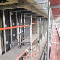 Montaż rusztowania przy elewacji budynku z widoczną warstwą izolacji i elementami mocującymi, narzędzia na podeście rusztowania.