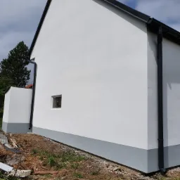 Boczny widok świeżo pomalowanej, białej elewacji budynku z szarym cokołem i czarnymi rynnami. Małe okno z białym obramowaniem. Na ziemi widoczne resztki materiałów budowlanych i gruzu.