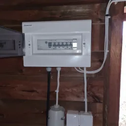 Biała skrzynka elektryczna z bezpiecznikami firmy Elektro-Plast zamontowana na drewnianej ścianie, z widocznymi przewodami i gniazdami.