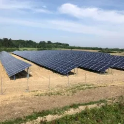 Rozległa farma paneli słonecznych na brązowej, piaszczystej ziemi, z zielonymi drzewami w tle pod błękitnym niebem z chmurami.