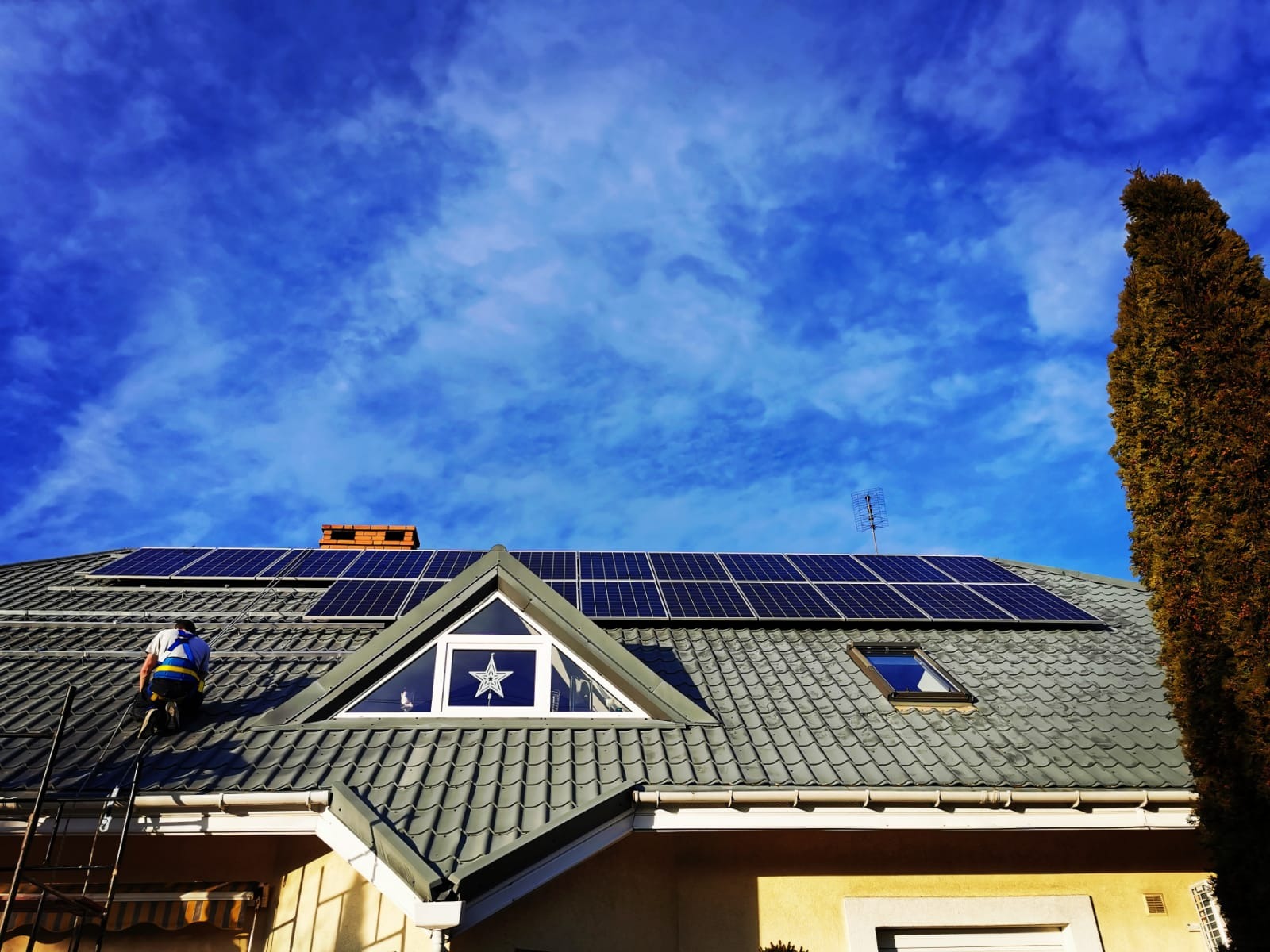 Montaż paneli słonecznych na dachu domu z szarą dachówką, pracownik na drabinie zabezpieczony uprzężą, błękitne niebo z chmurami w tle.