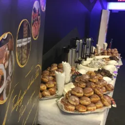 St&oacute;ł z białym obrusem, na kt&oacute;rym ułożone są tace z pączkami, ciasteczkami i wafle, obok stoją metalowe dystrybutory do kawy, w tle czarno-granatowa ściana z dekoracjami i napisem.