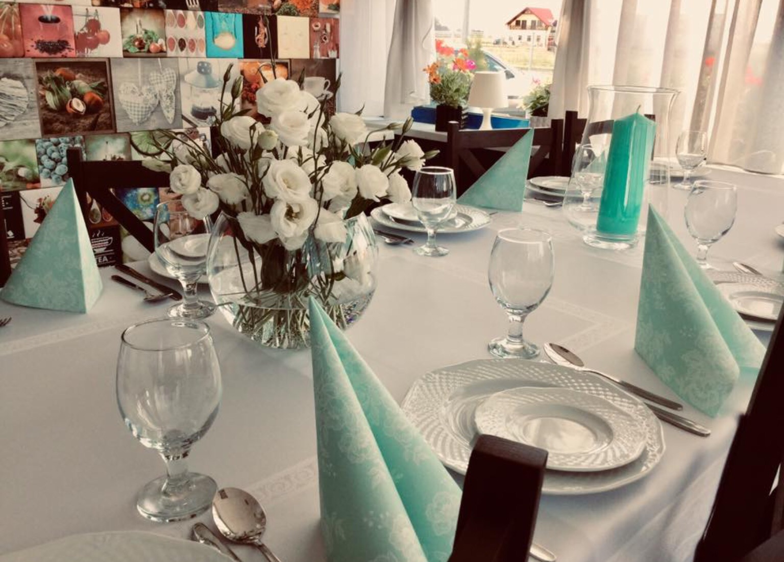 Uroczysty stół nakryty białym obrusem, dekorowany bukietem białych róż w szklanym wazonie, turkusowymi serwetkami w kształcie stożków, elegancką zastawą i wysoką, turkusową świecą.