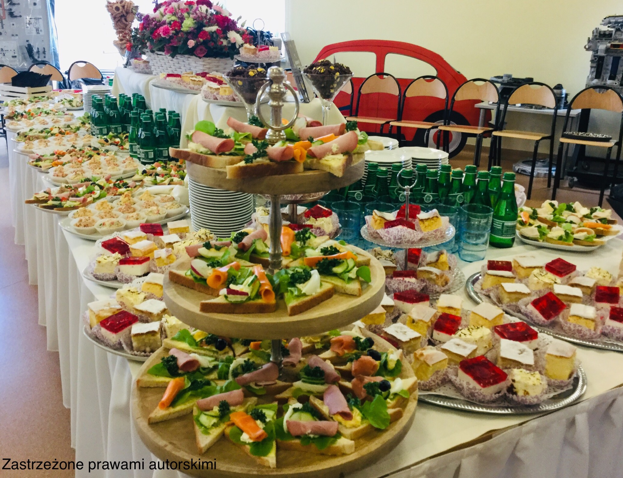 Bogato zastawiony stół z przekąskami: kanapki na drewnianym etażerze, koreczki, ciasta z galaretką, napoje w zielonych butelkach oraz dekoracje kwiatowe w tle.