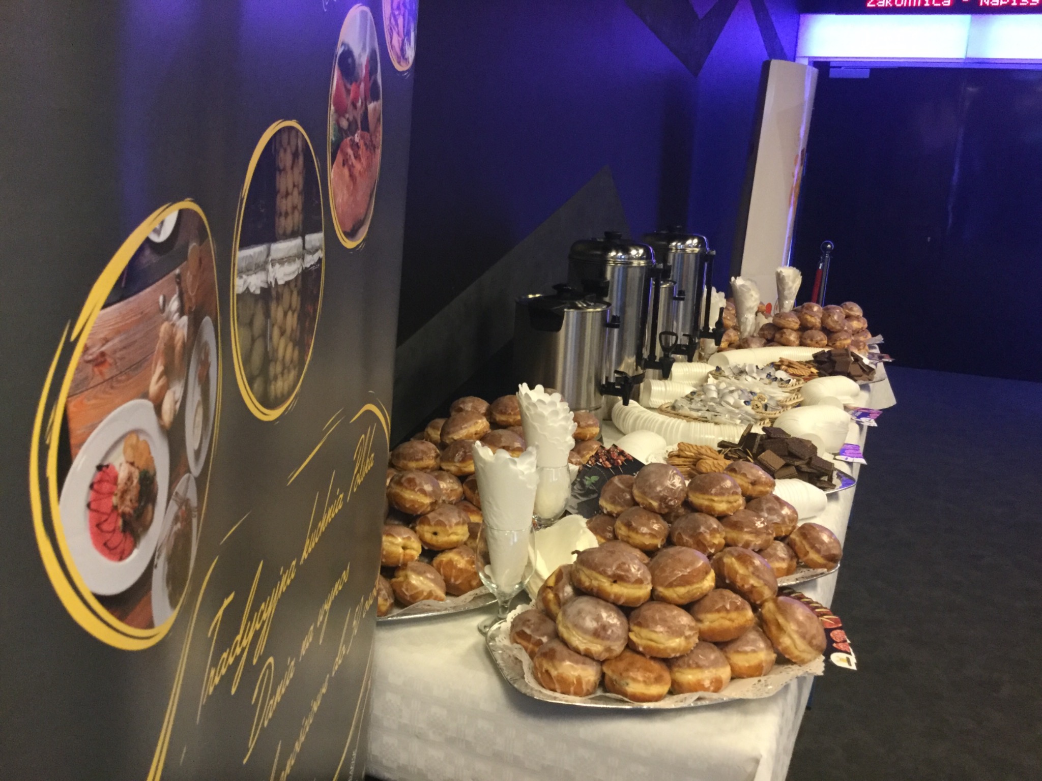 Stół z białym obrusem, na którym ułożone są tace z pączkami, ciasteczkami i wafle, obok stoją metalowe dystrybutory do kawy, w tle czarno-granatowa ściana z dekoracjami i napisem.