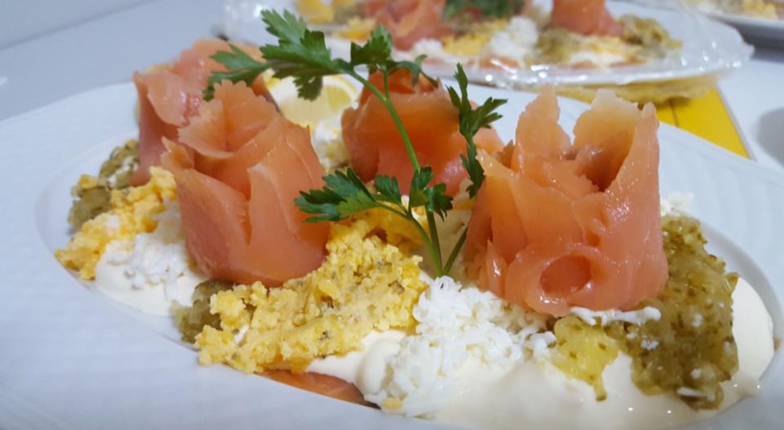 Elegancko podane danie z łososia uformowanego w kształt róży, z dekoracją z zielonej pietruszki, jajka na twardo, białego sosu i zielonego pesto na białym talerzu.