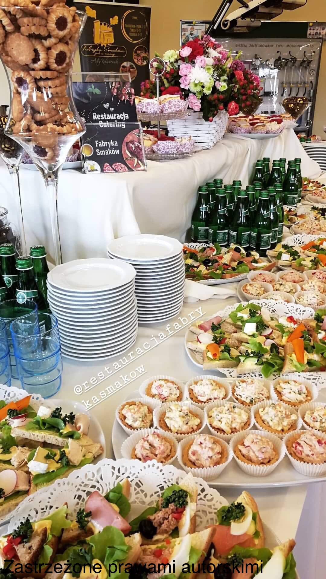 Stół cateringowy z przekąskami, tartaletkami, kanapkami, butelkami wody i ciasteczkami, aranżacja z kwiatami w tle, widoczny szyld reklamowy 'Restauracja Catering Fabryka Smaków'.