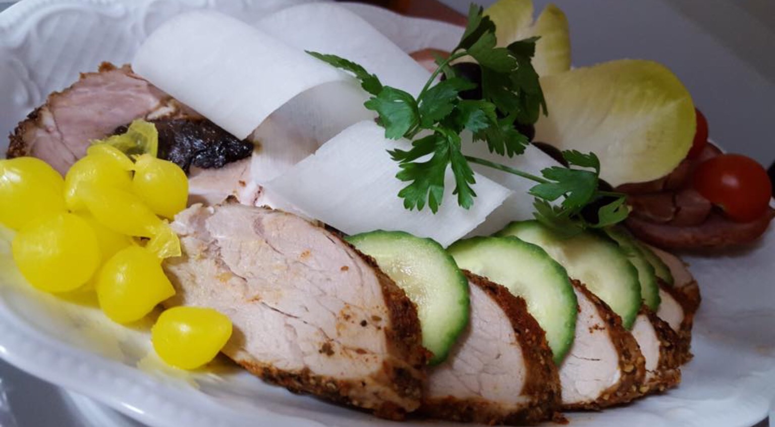 Elegancko ułożona na białym talerzu kompozycja z plastrów pieczonego mięsa, zielonego ogórka, żółtych marynowanych kulek, białych rulonów i zielonej pietruszki.