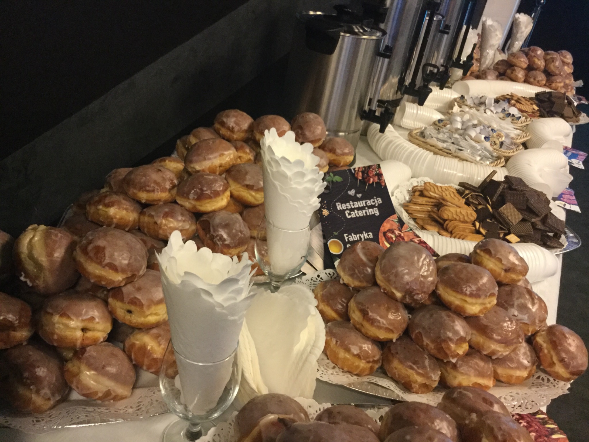 Stół z pączkami z lukrem, ciasteczkami i wafle czekoladowymi, dodatkowo srebrne termosy i białe kubki na tle ciemnej ściany.