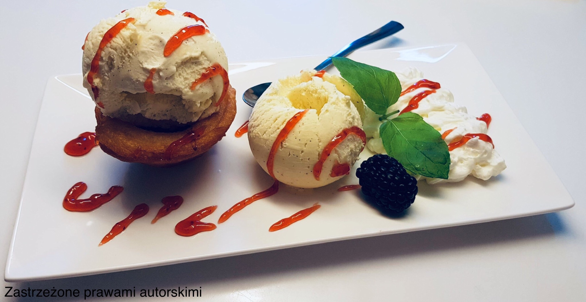 Elegancko podany deser: gałka lodów waniliowych na pączku, udekorowana sosem truskawkowym, bitą śmietaną, liśćmi mięty i jeżyną na białym talerzu.
