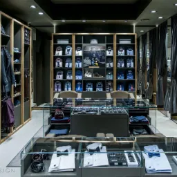 Ekskluzywny salon odzieżowy z męską kolekcją: koszule, krawaty, buty, akcesoria, prezentowane w szklanych gablotach i na półkach w zabudowie, z przymierzalniami zasłoniętymi szarymi kotarami w tle.