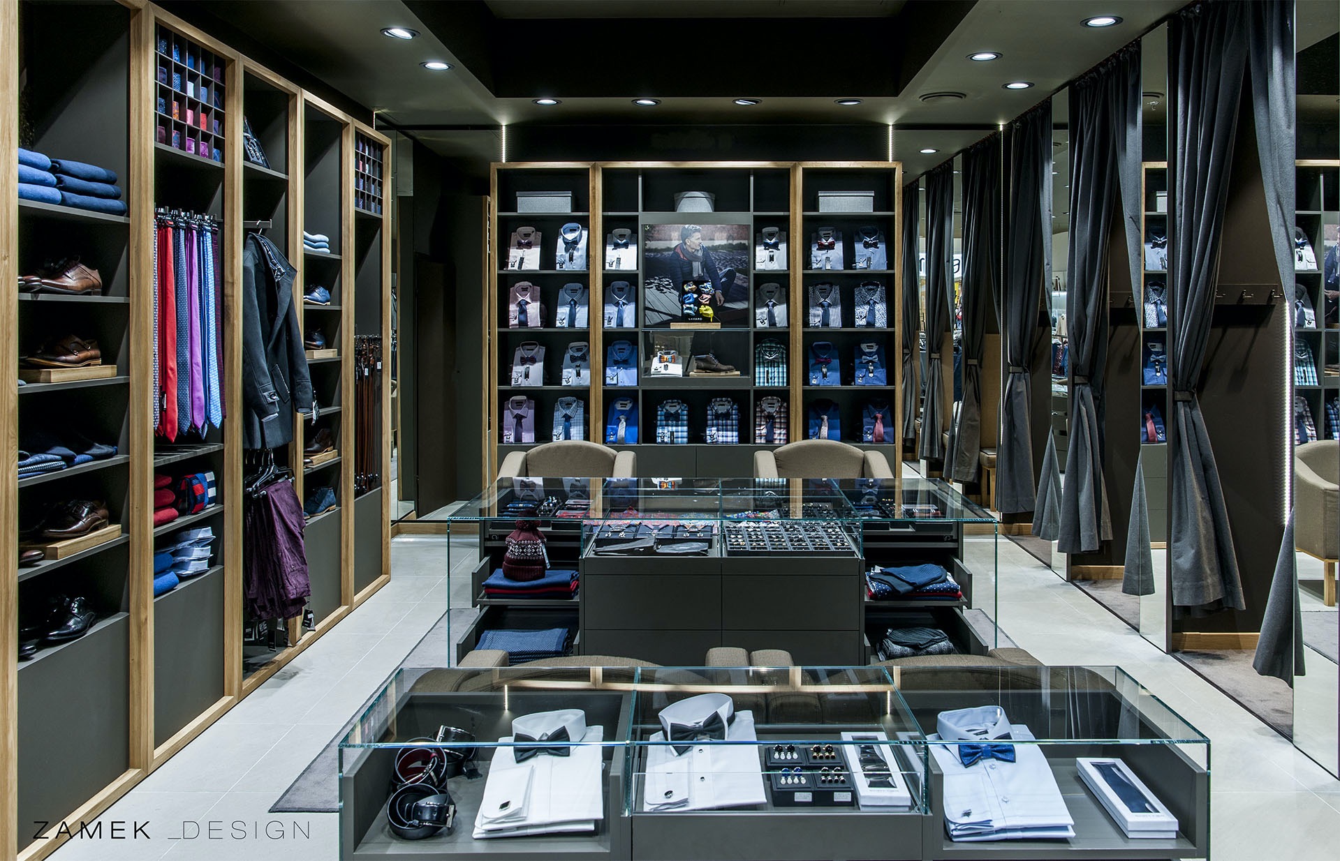 Ekskluzywny salon odzieżowy z męską kolekcją: koszule, krawaty, buty, akcesoria, prezentowane w szklanych gablotach i na półkach w zabudowie, z przymierzalniami zasłoniętymi szarymi kotarami w tle.