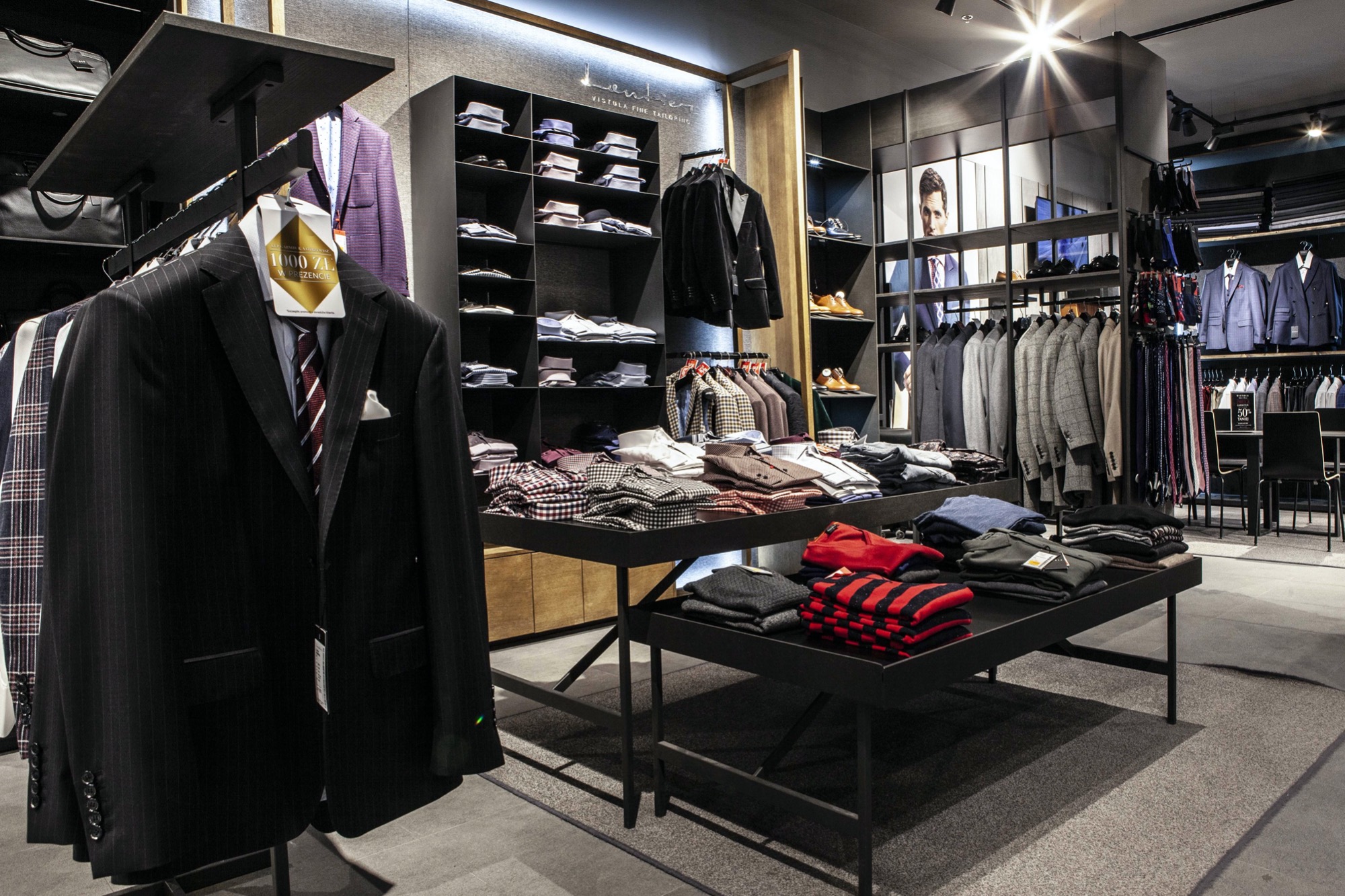 Wnętrze eleganckiego salonu odzieżowego z garniturami, koszulami i dodatkami, oświetlone punktowo, z minimalistycznymi czarnymi regałami i stołami ekspozycyjnymi.