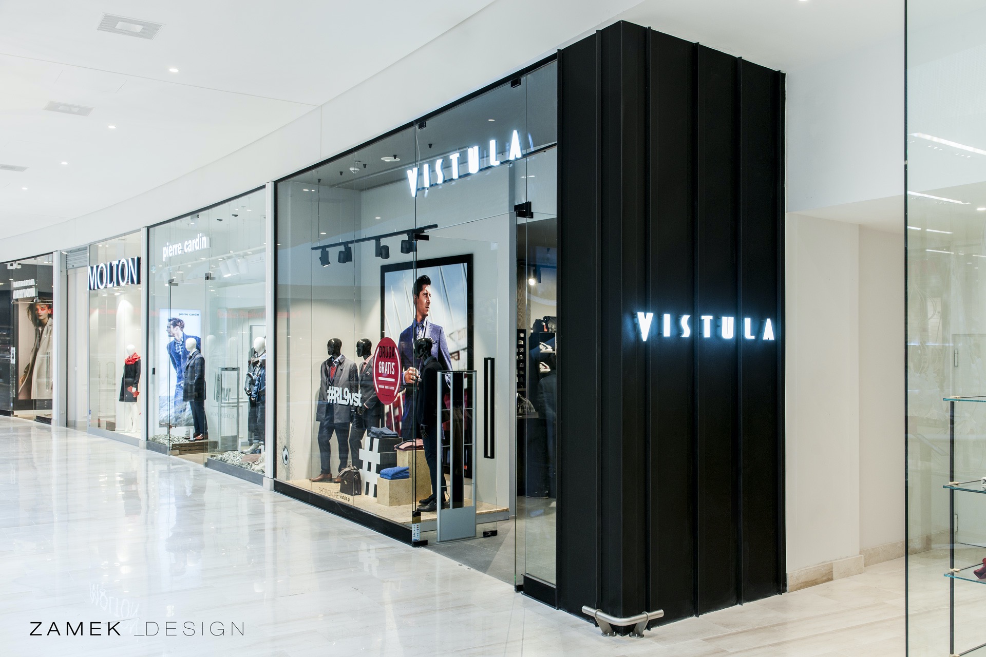 Elegancki wystrój sklepu odzieżowego Vistula z przeszkloną fasadą i podświetlanym logo na czarnym, żebrowanym obramowaniu, widoczne manekiny w garniturach i plakaty reklamowe wewnątrz.