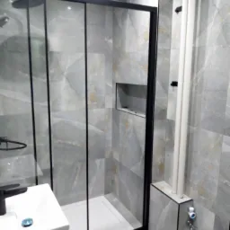 Wykończona łazienka z kabiną prysznicową typu walk-in, szare płytki imitujące marmur, czarna armatura i wnęka w ścianie. Widoczne pionowe rury przy ścianie.