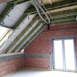 Poddasze w trakcie prac: widoczne drewniane krokwie, izolacja, instalacje wentylacyjne i okno. Ściany z czerwonej cegły. Etap przed wykończeniem.