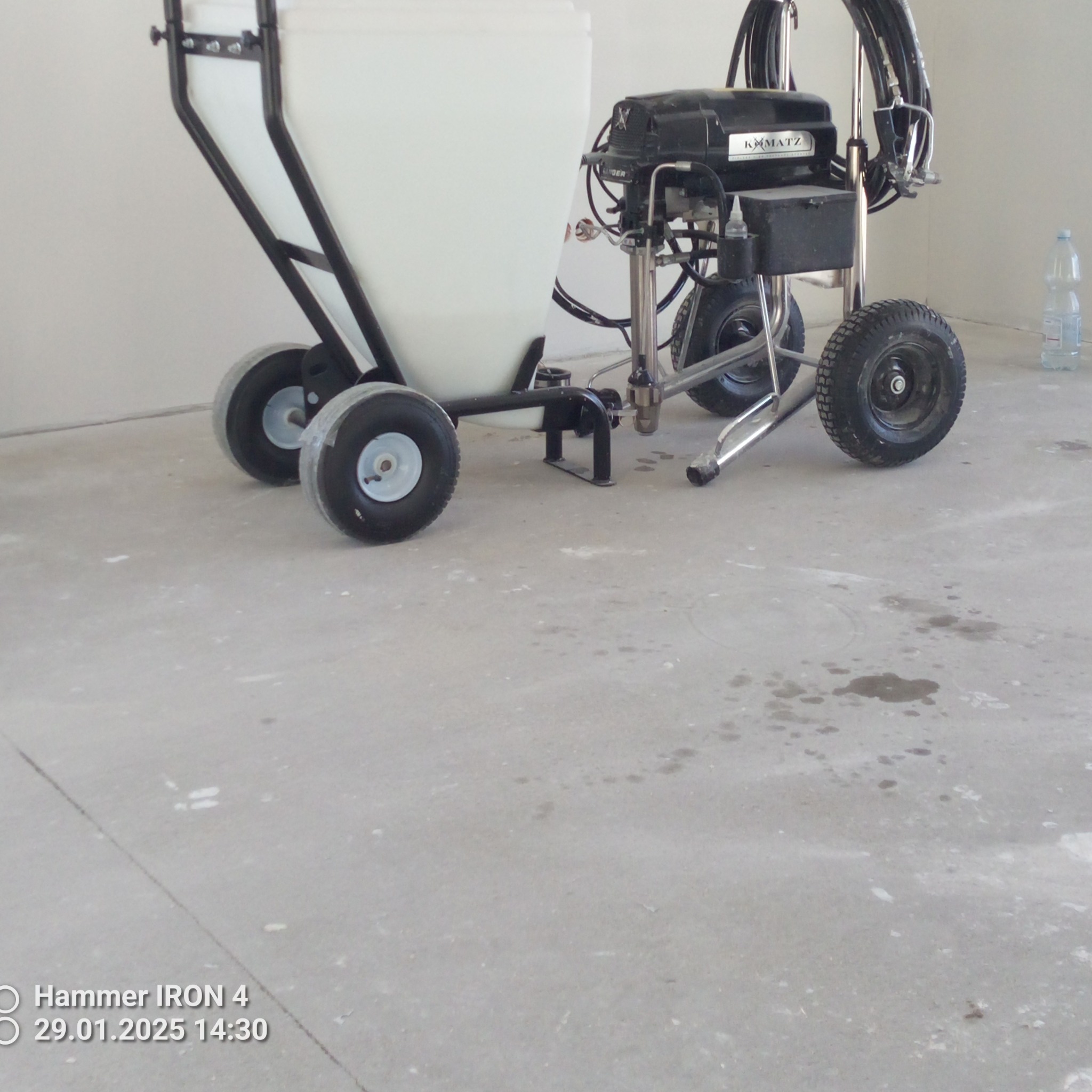 Profesjonalny agregat malarski Komatz na kółkach z dużym zbiornikiem, stojący na betonowej posadzce z widocznymi śladami farby i butelką wody w tle.