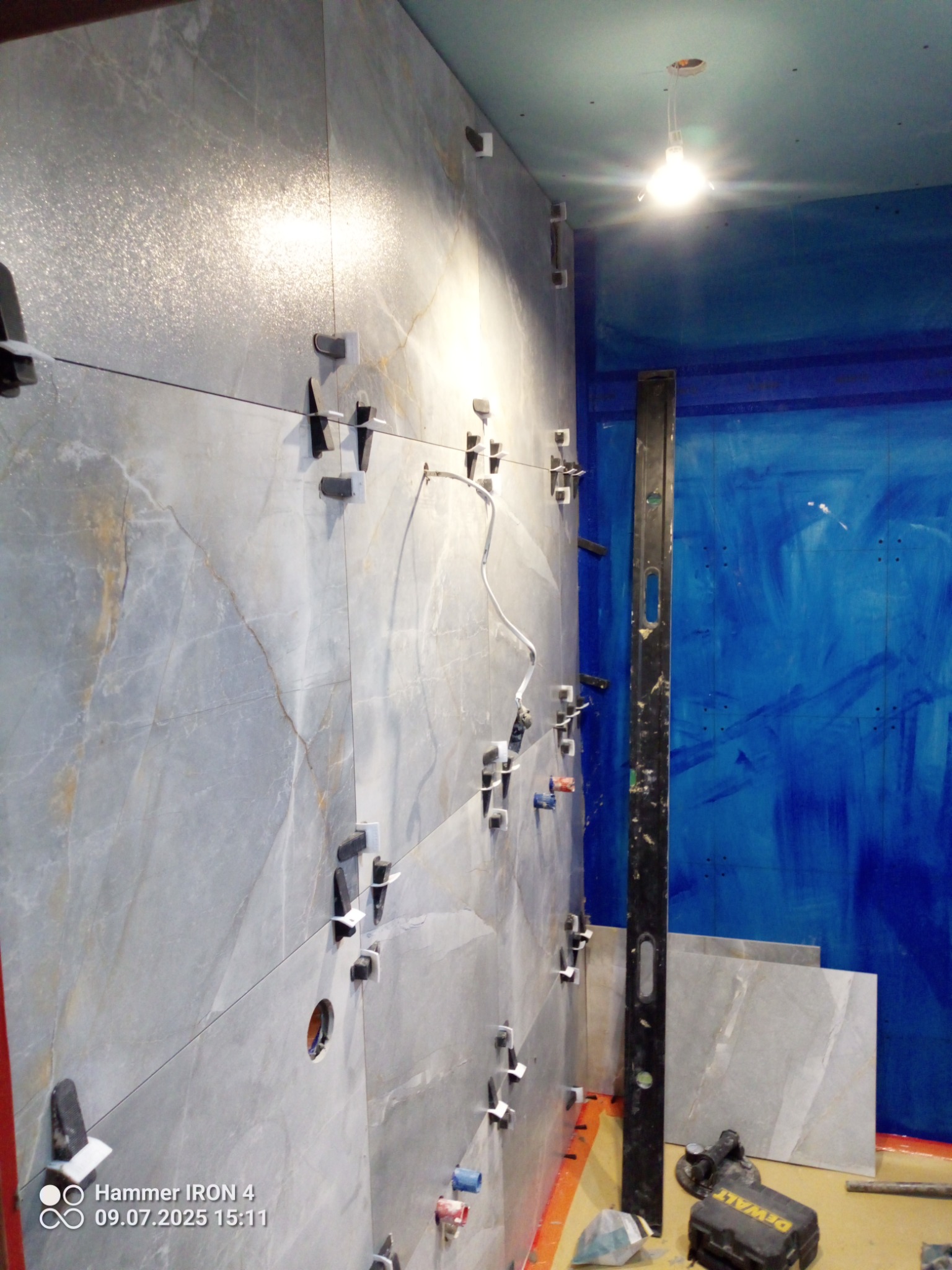 Układanie płytek imitujących marmur na ścianie z użyciem klipsów poziomujących. Widoczny poziom i narzędzia. Intensywny niebieski kolor ściany w tle.