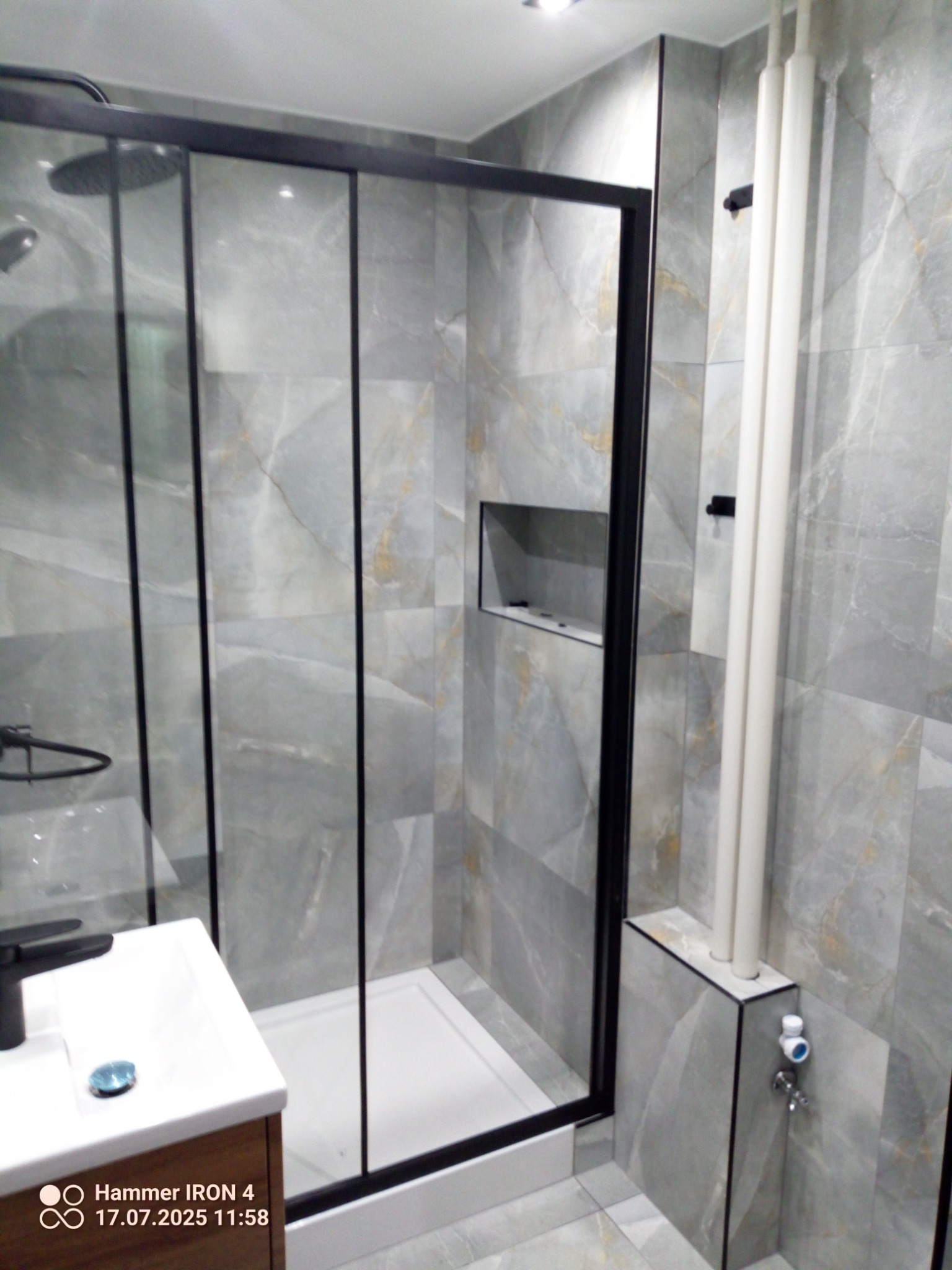 Wykończona łazienka z kabiną prysznicową typu walk-in, szare płytki imitujące marmur, czarna armatura i wnęka w ścianie. Widoczne pionowe rury przy ścianie.